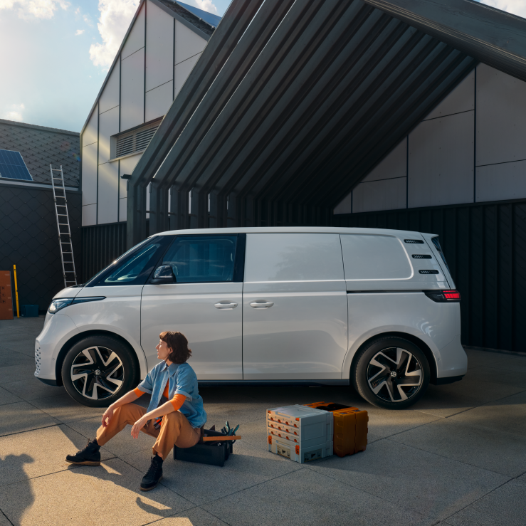 Ein weißer Volkswagen ID. Buzz Cargo vor einer Werkshalle, im Vordergrund sind ein Mann und eine Frau zu sehen