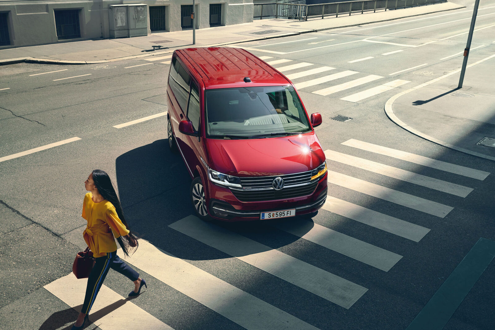 Një Multivan 6.1 i kuq ecën në një rrugë mbi vijat e bardha. Një grua në bluzë të verdhë përshkon rrugën.