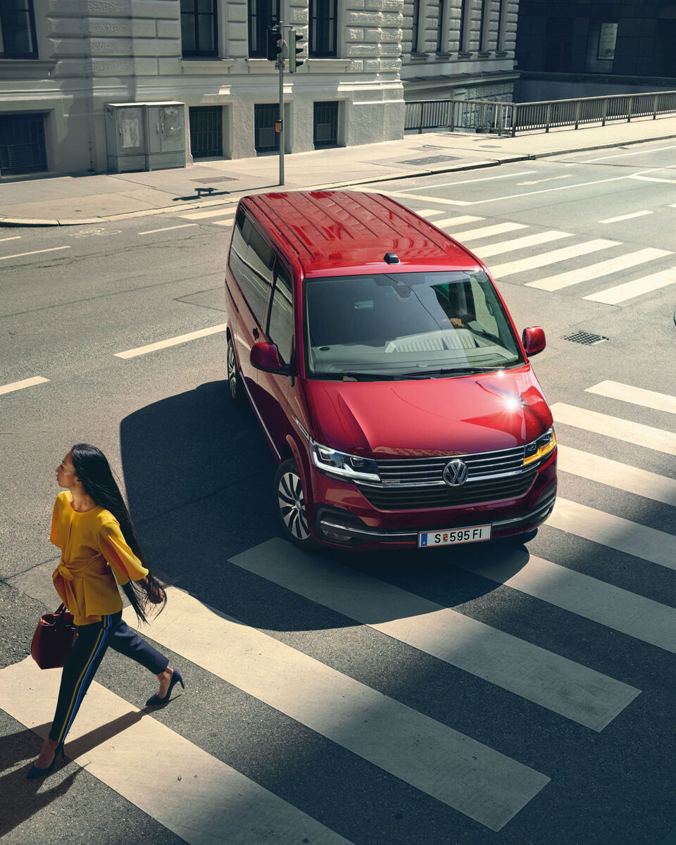 Një Multivan 6.1 i kuq ecën në një rrugë mbi vijat e bardha. Një grua në bluzë të verdhë përshkon rrugën.